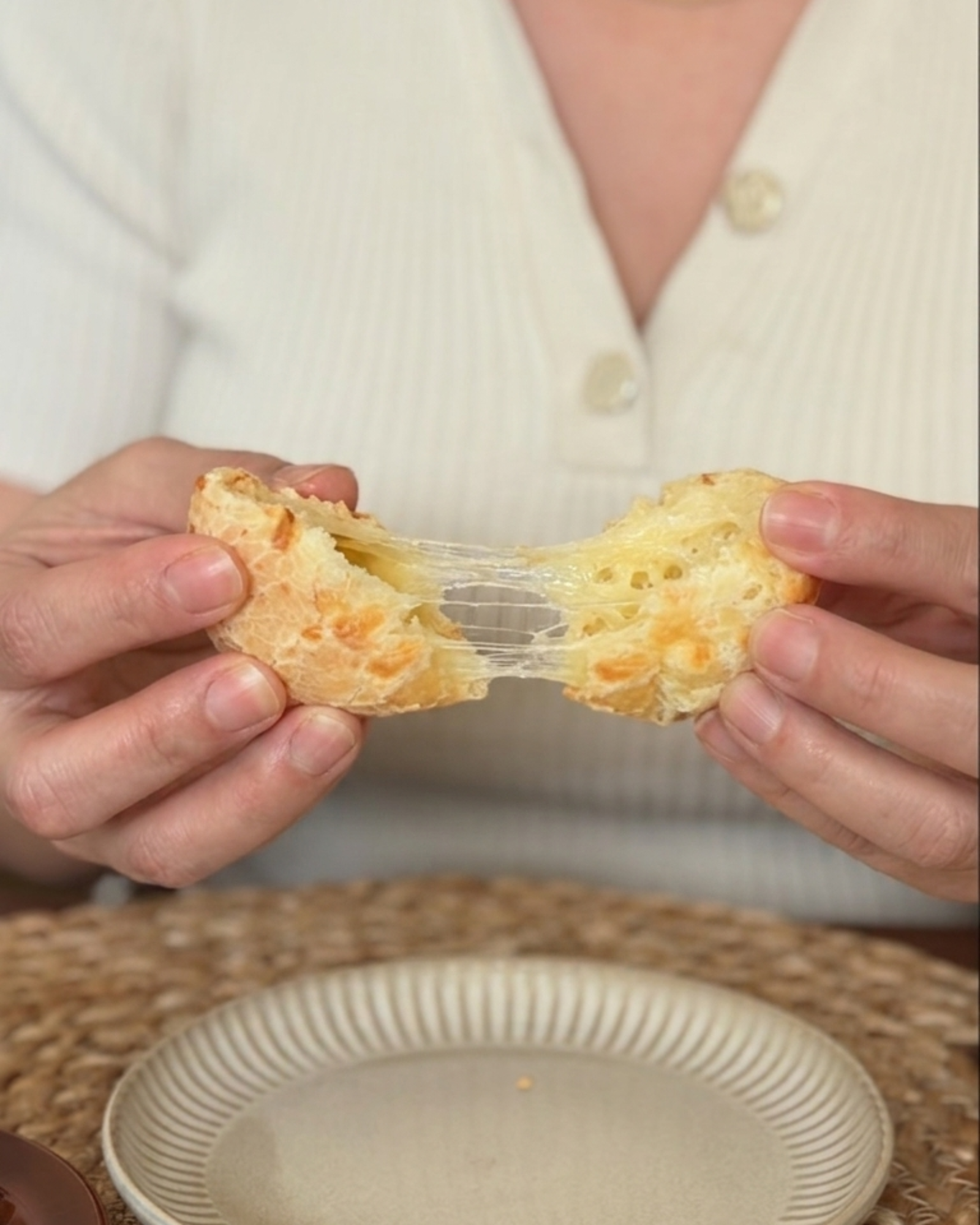Person holding a cheesy pão de queijo with a plate on a rattan mat.