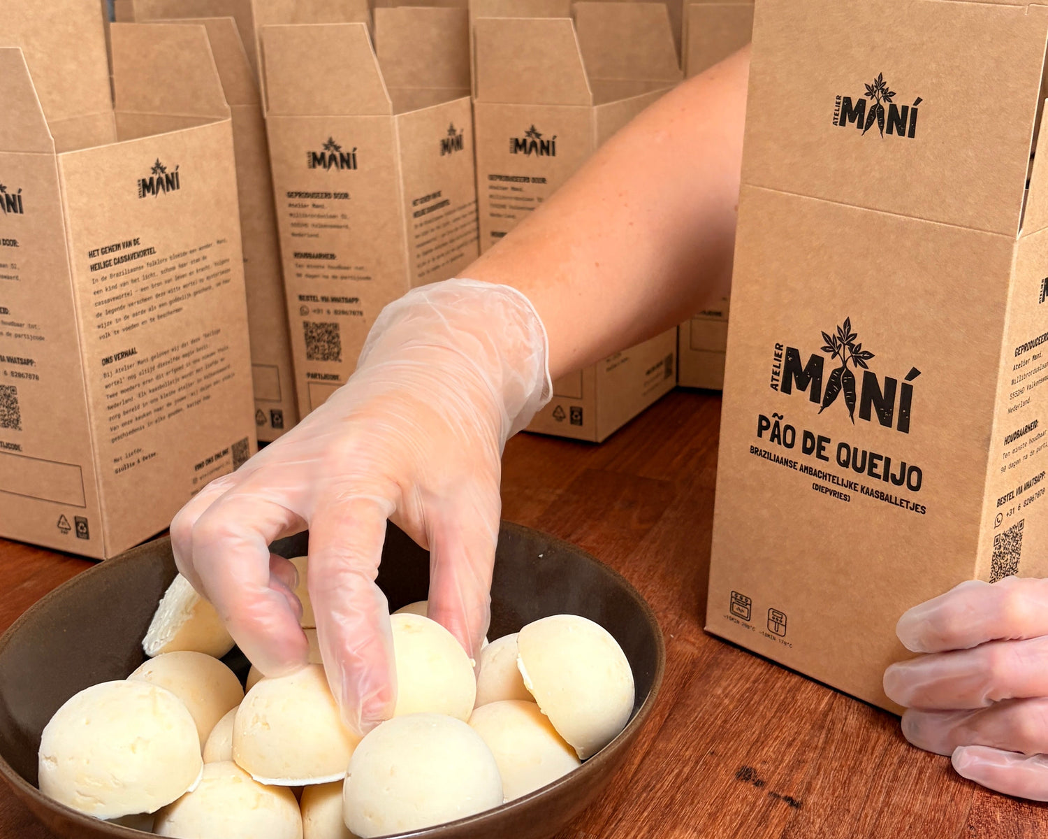 Person handling pão de queijo Maní branded boxes on a wooden table.
